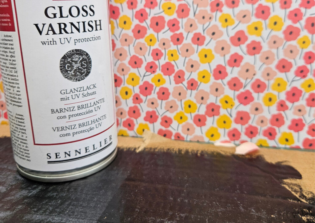 Close-up of a white varnish can labeled 'GLOSS VARNISH with UV protection' on a dark wooden surface, with bright floral wallpaper in the background.