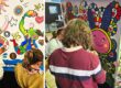 Group of people painting a large, colorful mural with joyful figures, sun and stars on a wall in an art workshop.