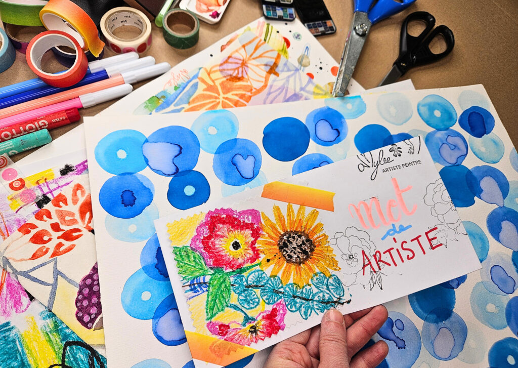 Assorted art supplies on a desk with colorful drawings and markers; a hand holds two small painted cards with sunflowers.