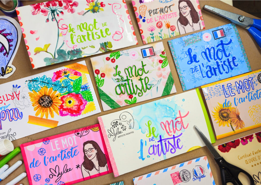 Colorful art postcards with 'Le mot de l'artiste' phrases spread on a craft table, tools like scissors and markers nearby.