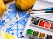 Watercolor paint set with colorful pans, two brushes, and lemons on a blue patterned tablecloth.
