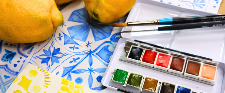 Watercolor paint set with colorful pans, two brushes, and lemons on a blue patterned tablecloth.