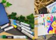 Blue notebook with a blue ribbon, surrounded by colorful markers and green ivy on a straw background; art supplies scene
