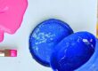 Pink paint spilling from a brush beside an open blue paint can on a light background with a canvas edge nearby.