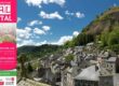 Bright pink festival poster on a mural announcing Festival Art en Cantal (Aug 8–9, 2026) with colorful drawings, over a town and hillside.