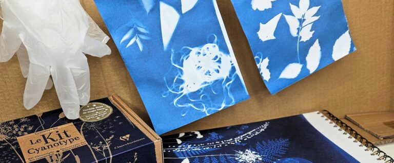 Gloved hand pointing at a cyanotype kit box labeled 'Le Kit Cyanotype' with blue botanical prints in the background.