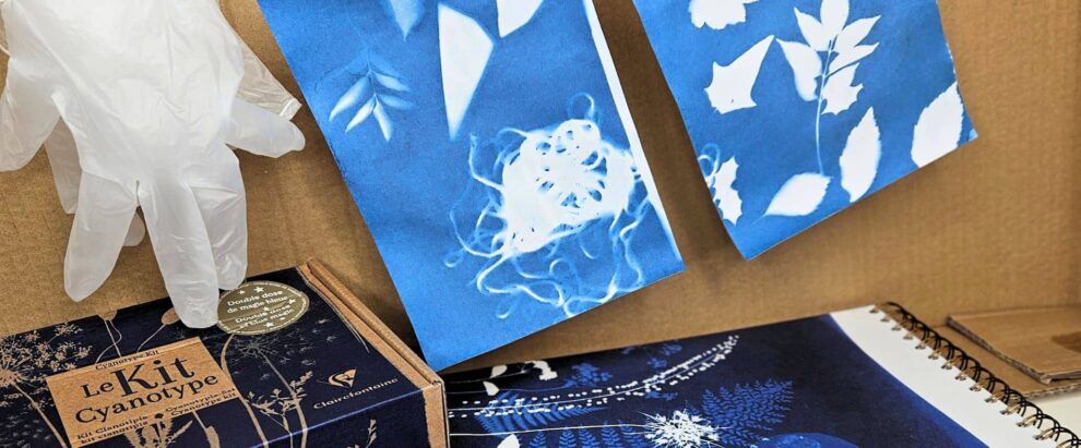 Gloved hand pointing at a cyanotype kit box labeled 'Le Kit Cyanotype' with blue botanical prints in the background.